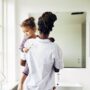 A Black mother holding her child near the sink of a clean white bathroom. They are both wearing pajama outfits.