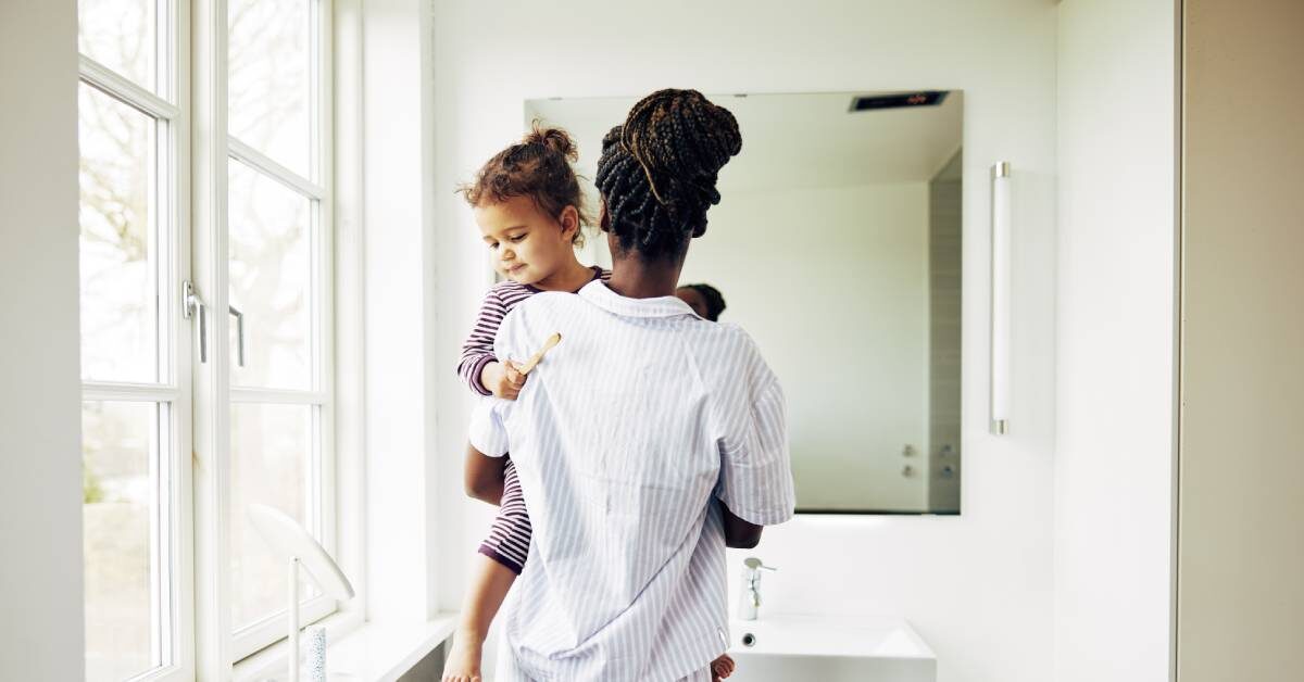 A Black mother holding her child near the sink of a clean white bathroom. They are both wearing pajama outfits.