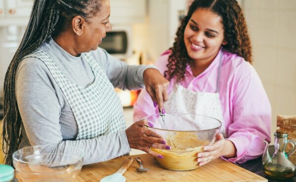 Two women wearing aprons mix batter in a glass bowl on a wooden countertop in a bright kitchen with various ingredients.