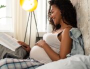 A black pregnant mother wearing a white tank and holding her hand over her stomach while lying in bed reading a book.