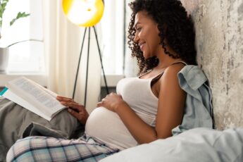 A black pregnant mother wearing a white tank and holding her hand over her stomach while lying in bed reading a book.