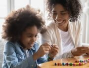 A woman and young girl smile as they add colorful beads to a string while sitting on a gray couch.