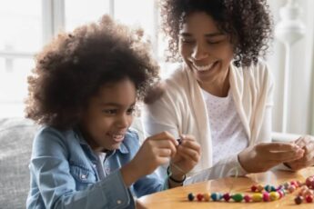 A woman and young girl smile as they add colorful beads to a string while sitting on a gray couch.