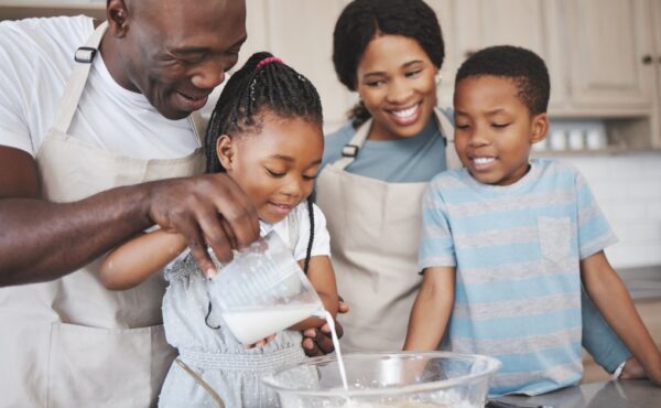 A black family is in the kitchen, bonding with each other. The father pours milk in the mix, and the daughter mixes.