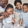 A black family is in the kitchen, bonding with each other. The father pours milk in the mix, and the daughter mixes.