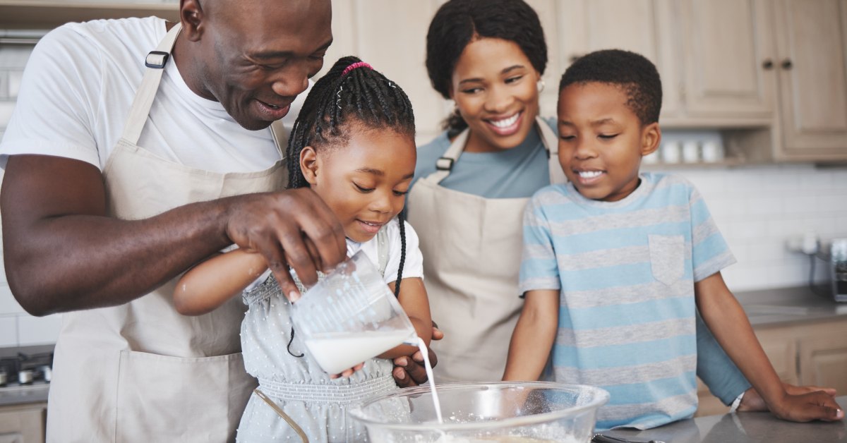 A black family is in the kitchen, bonding with each other. The father pours milk in the mix, and the daughter mixes.