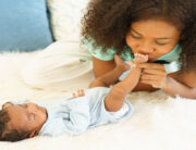 A close-up showing a woman wearing a green shirt kissing the foot of a small baby on top of a white rug.