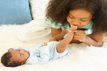 A close-up showing a woman wearing a green shirt kissing the foot of a small baby on top of a white rug.