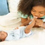 A close-up showing a woman wearing a green shirt kissing the foot of a small baby on top of a white rug.