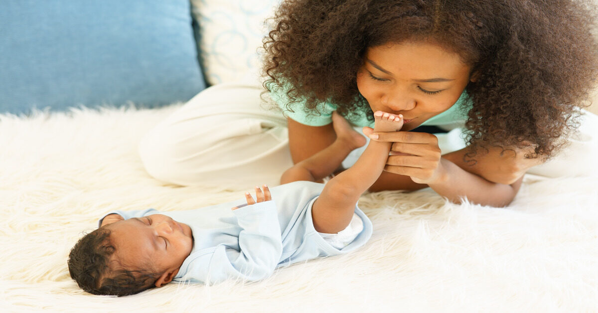 A close-up showing a woman wearing a green shirt kissing the foot of a small baby on top of a white rug.
