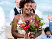 A smiling bride stands on the beach holding a colorful bouquet. She's surrounded by clapping people in formal wear.