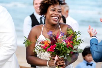 A smiling bride stands on the beach holding a colorful bouquet. She's surrounded by clapping people in formal wear.
