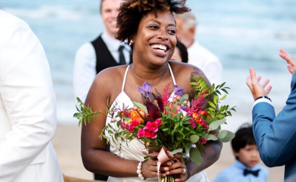 A smiling bride stands on the beach holding a colorful bouquet. She's surrounded by clapping people in formal wear.