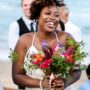 A smiling bride stands on the beach holding a colorful bouquet. She's surrounded by clapping people in formal wear.
