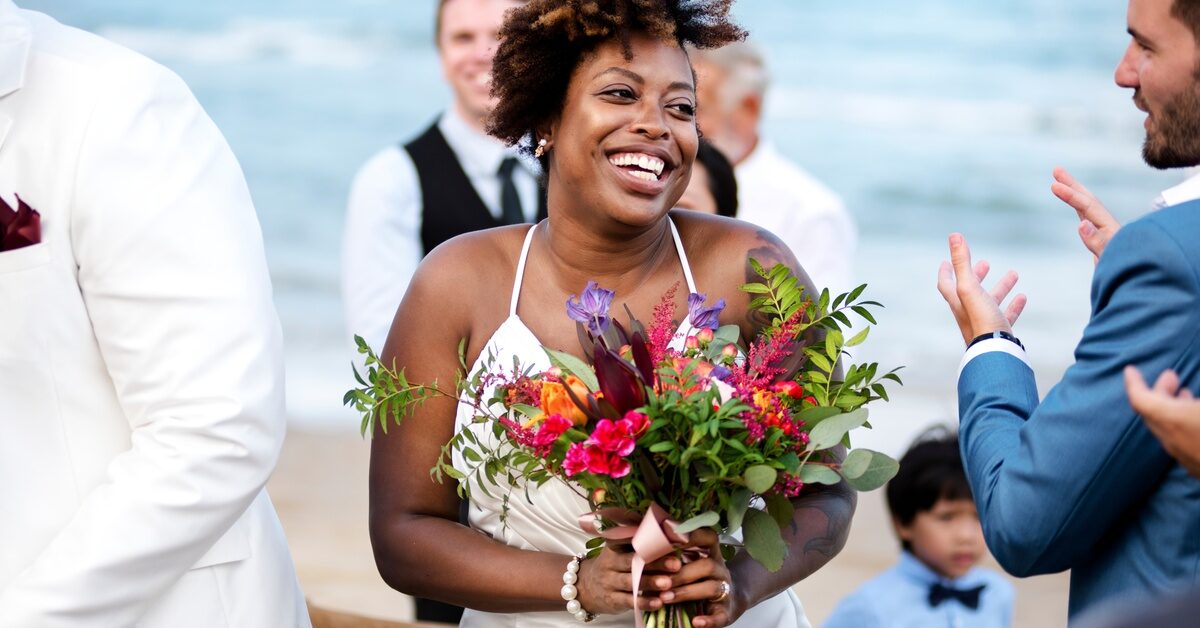A smiling bride stands on the beach holding a colorful bouquet. She's surrounded by clapping people in formal wear.
