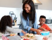 A woman stands in a kitchen with her two children while packing sandwiches and fruit into lunch boxes.