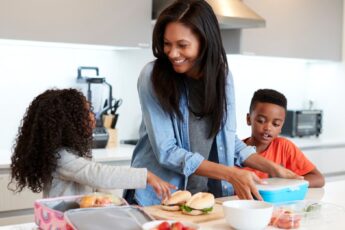 A woman stands in a kitchen with her two children while packing sandwiches and fruit into lunch boxes.