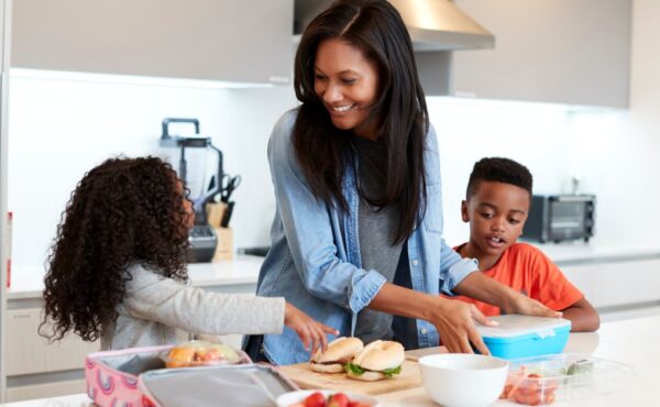 A woman stands in a kitchen with her two children while packing sandwiches and fruit into lunch boxes.