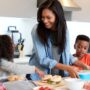 A woman stands in a kitchen with her two children while packing sandwiches and fruit into lunch boxes.