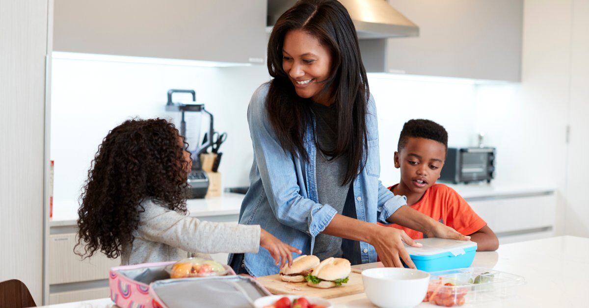 A woman stands in a kitchen with her two children while packing sandwiches and fruit into lunch boxes.