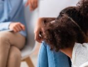 A back view of a child sitting with her head on her knees as she talks with an adult sitting across from her.