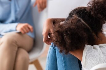 A back view of a child sitting with her head on her knees as she talks with an adult sitting across from her.
