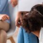 A back view of a child sitting with her head on her knees as she talks with an adult sitting across from her.