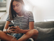 A Black teenaged girl sitting on her family's dark gray couch. She's holding a smartphone with wired ear buds in.