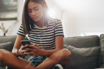 A Black teenaged girl sitting on her family's dark gray couch. She's holding a smartphone with wired ear buds in.