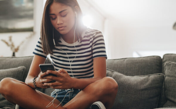A Black teenaged girl sitting on her family's dark gray couch. She's holding a smartphone with wired ear buds in.