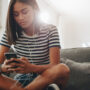 A Black teenaged girl sitting on her family's dark gray couch. She's holding a smartphone with wired ear buds in.