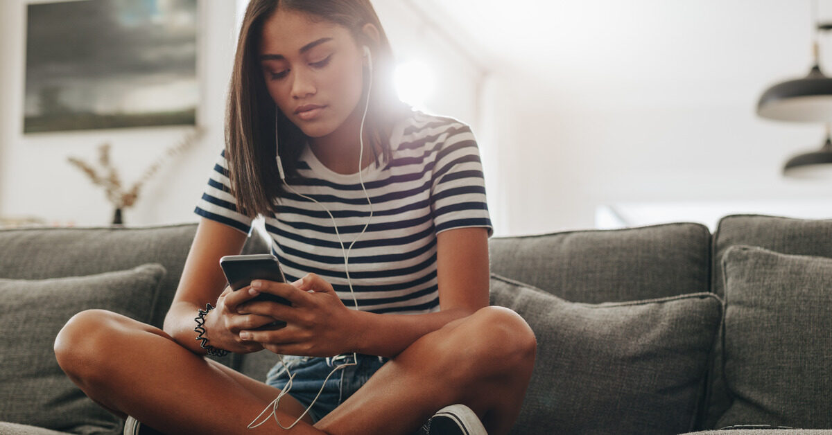 A Black teenaged girl sitting on her family's dark gray couch. She's holding a smartphone with wired ear buds in.