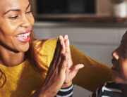 A Black mother and her daughter sitting on the front steps at home high-fiving each other with smiles on their faces.