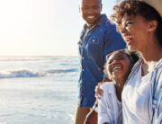 A Black family walks on a beach together. The mom, dad, and child all have smiles on their faces.