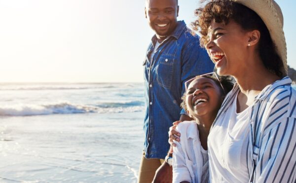 A Black family walks on a beach together. The mom, dad, and child all have smiles on their faces.