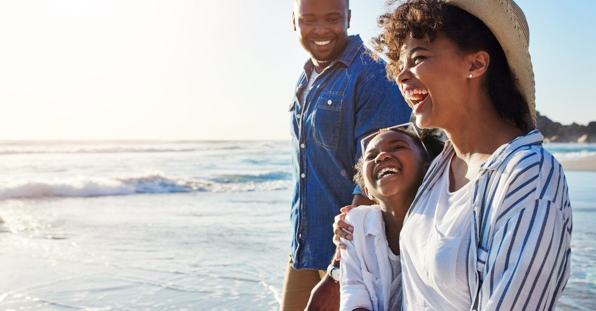 A Black family walks on a beach together. The mom, dad, and child all have smiles on their faces.