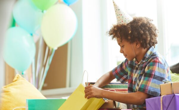 A young Black boy wearing a birthday party hat and opening birthday gifts. He's smirking as he opens a yellow gift bag.