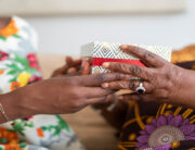 A Black adult daughter giving her grandmother a special box with a floral print. The grandmother is receiving the box.