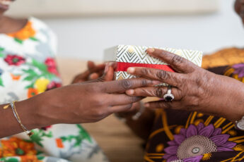 A Black adult daughter giving her grandmother a special box with a floral print. The grandmother is receiving the box.