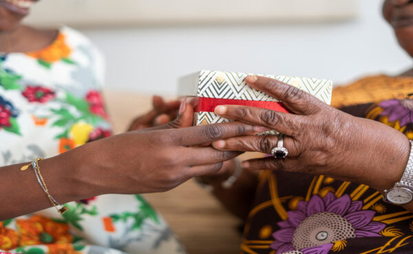 A Black adult daughter giving her grandmother a special box with a floral print. The grandmother is receiving the box.