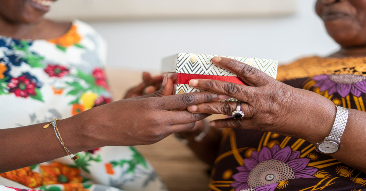 A Black adult daughter giving her grandmother a special box with a floral print. The grandmother is receiving the box.