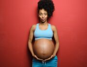 A black woman making a funny face while she poses in front of a red background. She is cradling her pregnant belly.
