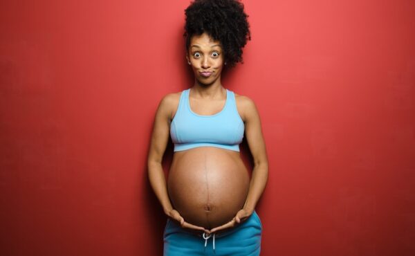 A black woman making a funny face while she poses in front of a red background. She is cradling her pregnant belly.