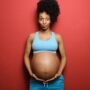 A black woman making a funny face while she poses in front of a red background. She is cradling her pregnant belly.