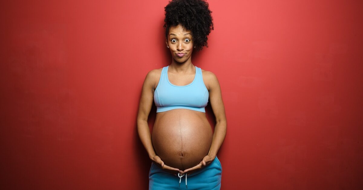 A black woman making a funny face while she poses in front of a red background. She is cradling her pregnant belly.