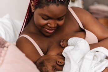 A Black mother on a bed smiles gently as she cradles her newborn and watches them breastfeed.