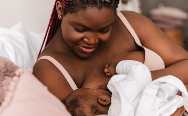 A Black mother on a bed smiles gently as she cradles her newborn and watches them breastfeed.
