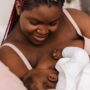 A Black mother on a bed smiles gently as she cradles her newborn and watches them breastfeed.