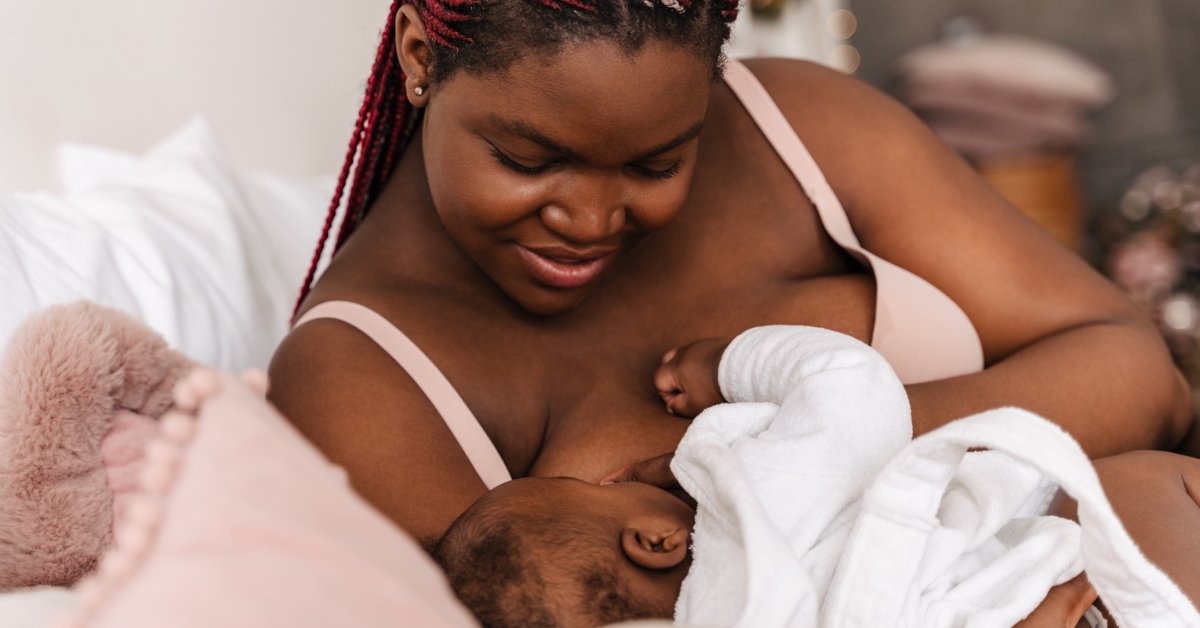 A Black mother on a bed smiles gently as she cradles her newborn and watches them breastfeed.