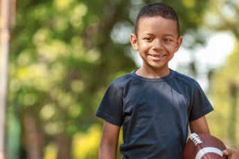 A smiling young boy wearing a black t-shirt stands in a park holding a football. Trees appear in the background.